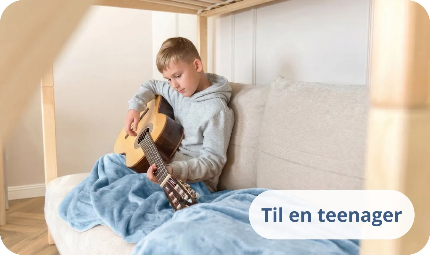 Teenager spiller akustisk guitar på en seng dækket af et blåt tæppe.